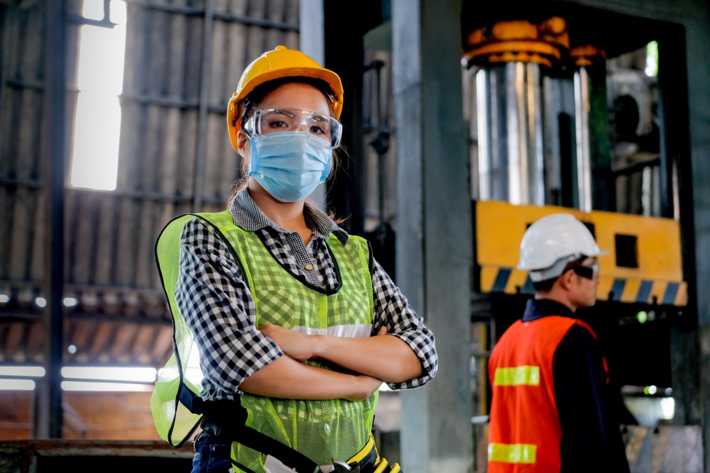 A woman wearing a face mask and construction helmet stands with arms crossed confidently.