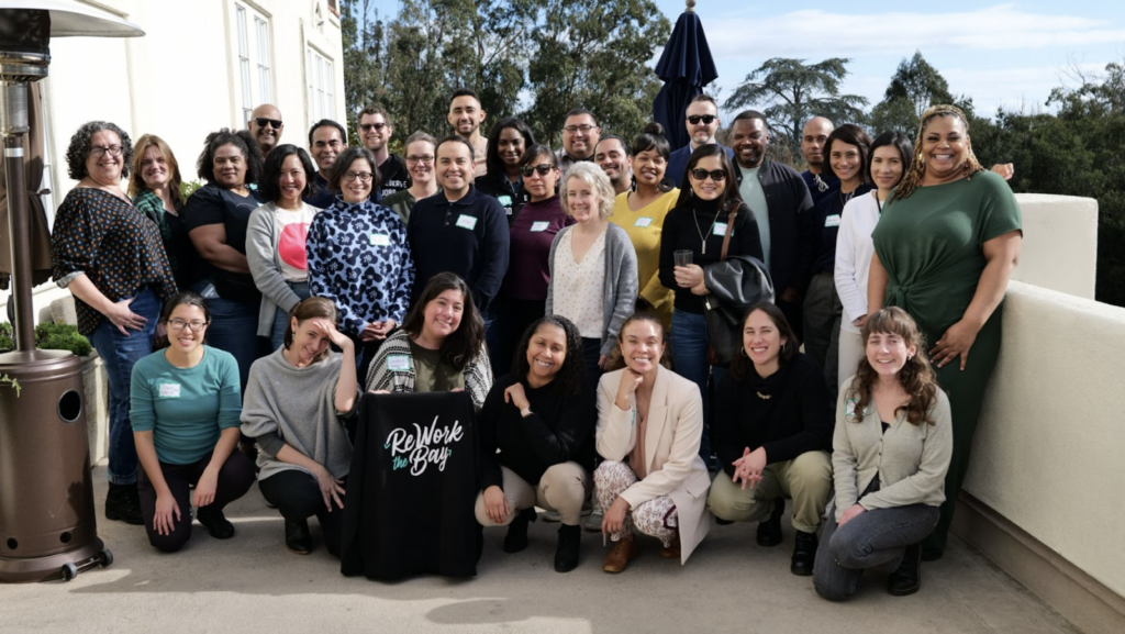 Photo of Rework The Bay team at a retreat, standing in the sun smiling. A woman in the front is holding a black sweatshirt with the organization's logo in white letters.